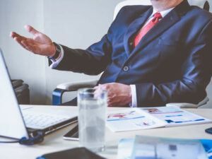 Business man in a navy blue suit sitting at his desk talking with someone in his office.