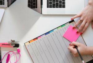 Woman's hands on an open planner and on a pink sticky note as she thinks and plans.